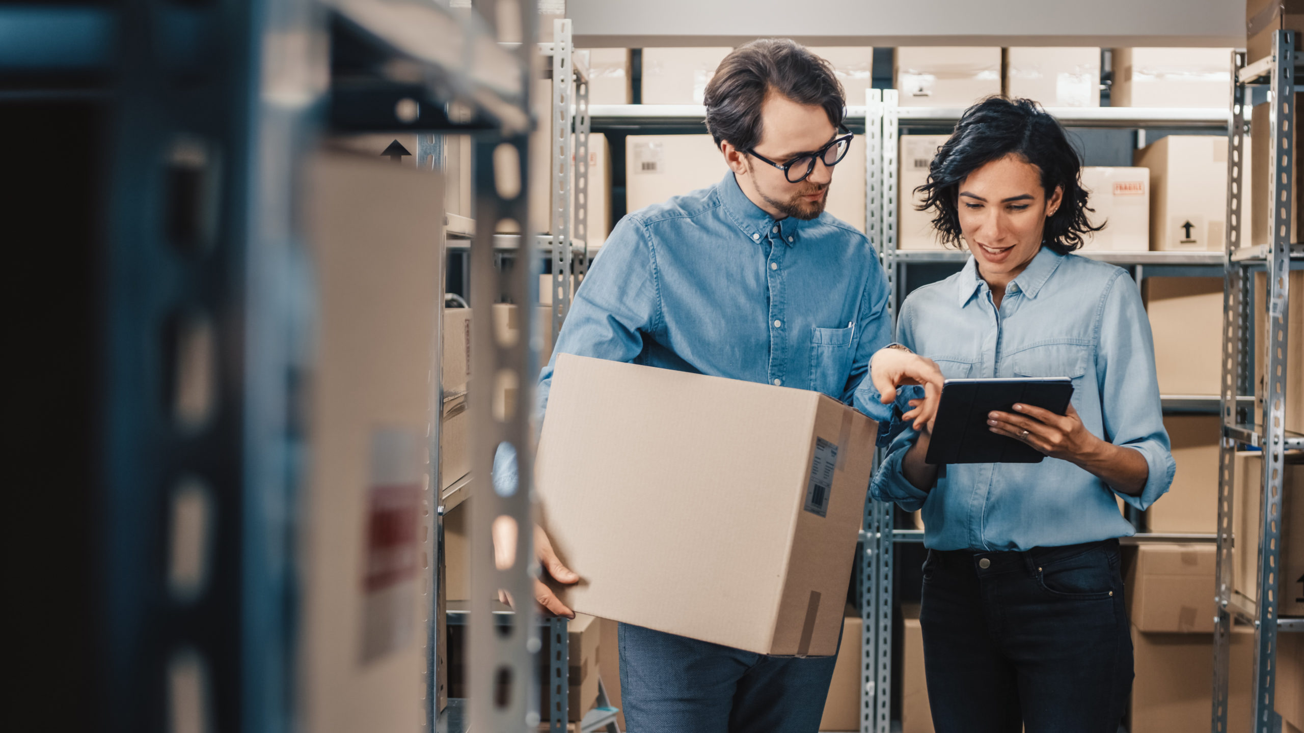 Female Inventory Manager Shows Digital Tablet Information to a Worker Holding Cardboard Box, They Talk and Do Work. In the Background Stock of Parcels with Products Ready for Shipment